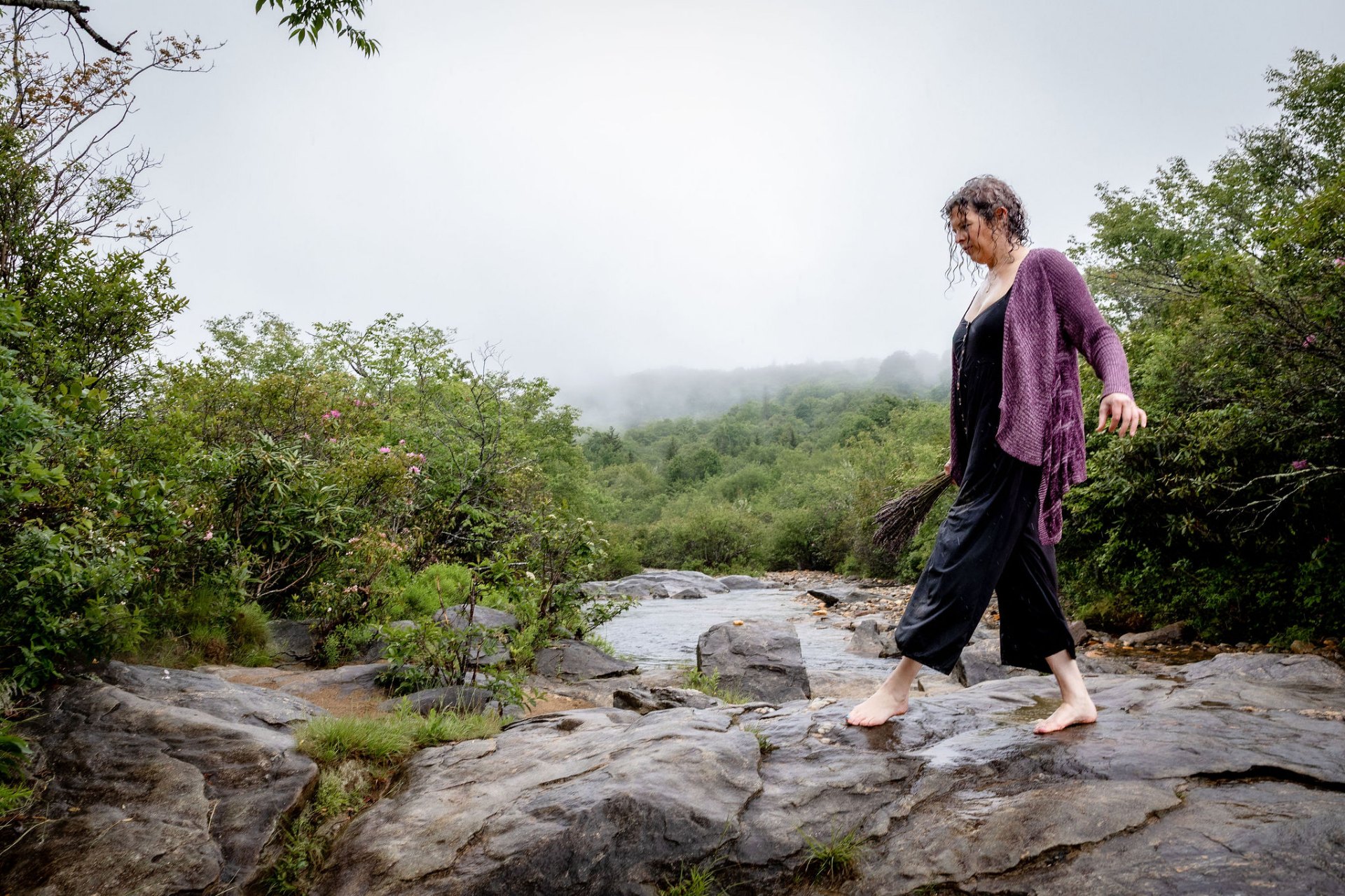 A woman in a dark dress and plum cardigan walks barefoot across wet stone at the edge of a misty mountain ridge, a small bundle of gathered branches in her hand