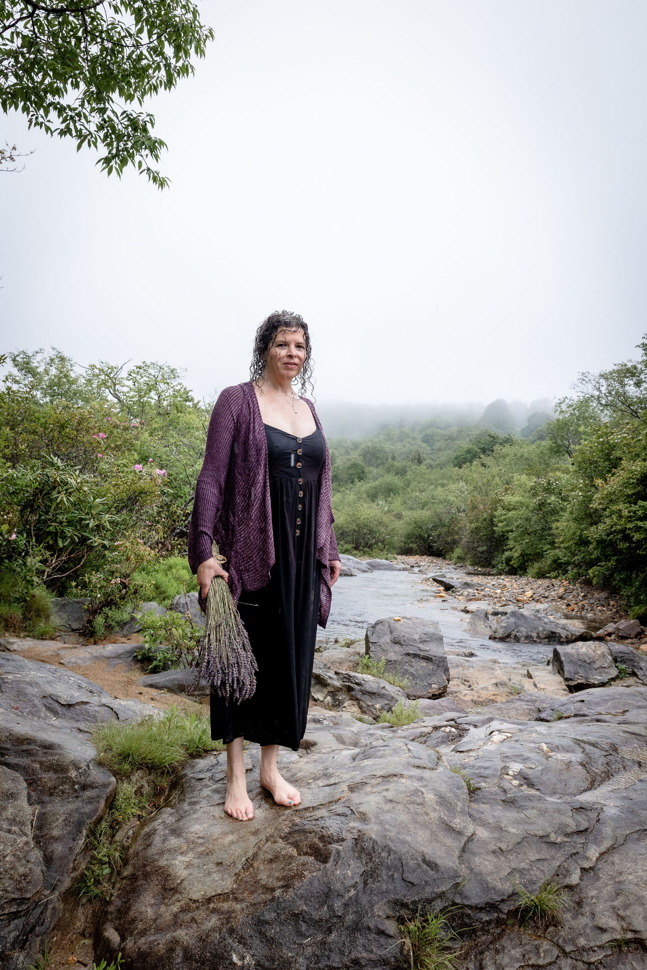 A woman stands barefoot on river stones in a misty stream, facing a threshold in the forest